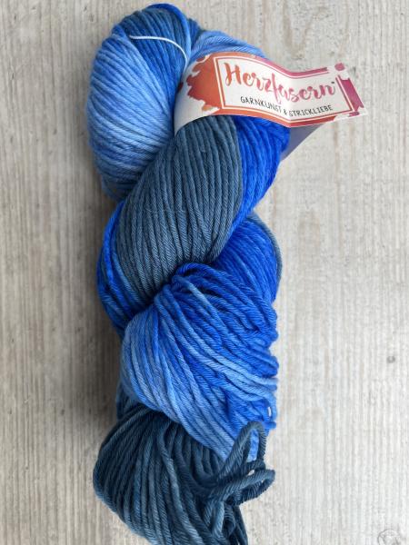 Handgefärbter Strang in Hellblau, Türkis und Mittelblau mit kleinen Farbsprenkelchen. Der Strang liegt auf hellem Holz, mit Herzfasern-Banderole.“